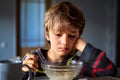Dissatisfied Caucasian young boy is having breakfast while sitting in front of transparent bowl of porridge in dimly lit