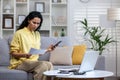 Dissatisfied and disappointed woman behind paper work sitting on sofa at home, Hispanic woman holding utility bills