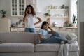 Dissatisfied European woman talking on phone standing in house behind little daughter sits on couch