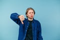 Dissatisfied guy in a shirt stands on a blue background and shows a dislike gesture with his fingers with a displeased face