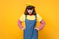 Dissatisfied sad girl teenager in french beret, denim sundress standing with arms akimbo isolated on yellow wall Royalty Free Stock Photo