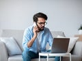 Busy Eastern Freelancer Guy Talking On Phone And Using Laptop At Home Royalty Free Stock Photo