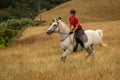 A distant shot of a beautiful happy smiling young woman dressed in a red polo shirt riding her white horse through long dried