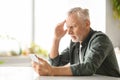 Distressed elderly man suffering headache while looking at smartphone at home