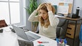 A distressed young woman grasping her head in an office setting with a laptop and workspace visible Royalty Free Stock Photo