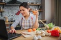 Disturbed woman sit on table in kitchen. She hold glass of wine and type on keyboard. Model look on laptop screen. She Royalty Free Stock Photo