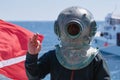 Diver wearing historical metallic helmet showing signal OK, diving flag and boat on the background