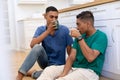 Diverse gay male couple sitting on floor in kitchen holding mugs and drinking Royalty Free Stock Photo