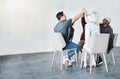 Diverse group of businesspeople giving each other a high five sitting on chairs in a meeting in an office at work. Happy Royalty Free Stock Photo