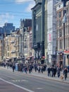 A diverse group of people walks in a lively hustle down a bustling city street flanked by soaring skyscrapers, under a