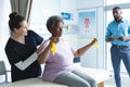 Diverse senior female patient exercising with band and female doctor advising in hospital room