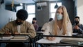 Diverse students in medical mask sit in classroom at lesson concentrated listen to lecture writing notes in notebook Royalty Free Stock Photo