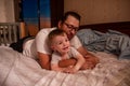 Diversity father with long hair, glasses hugs little son. Young man fooling around with boy on bed Royalty Free Stock Photo