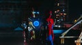 Two cosplayers stand together in images of a character Spider-Man and Iron Man against cityscape and night lights