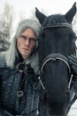 Portrait of cosplayer in image of a character Geralt of Rivia from the game or film The Witcher next to a horse in forest