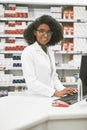 We do bookings as well. Portrait of a cheerful young female pharmacist typing on a computer while looking at the camera. Royalty Free Stock Photo