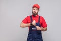 We do ontime. Young confident handyman with beard in blue overall and red t-shirt standing and showing time on his wrist watch wi Royalty Free Stock Photo