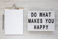 `Do what makes you happy` words on a lightbox, clipboard with blank sheet of paper on a white wooden background, top view. Royalty Free Stock Photo