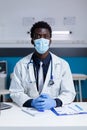 Doctor of african american ethnicity sitting at desk with face mask Royalty Free Stock Photo