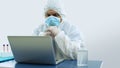 Doctor with blue mask and bioprotective suit working with his laptop in his office with his head resting on his hands with Royalty Free Stock Photo