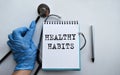 A doctor in blue medical gloves holds a notebook with the inscription HEALTHY HABITS, on the background of a light table, along Royalty Free Stock Photo