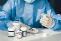 Doctor in a blue surgical gown and mask holds in his hand a medicine bottle. Many pills and drugs on white table, healthcare