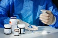Doctor in a blue surgical gown and mask holds in his hand a medicine bottle. Many pills and drugs on white table, healthcare