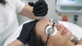 The doctor cosmetologist in a dressing gown and gloves performs laser procedures on the face of a female patient close