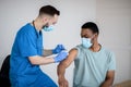 Doctor in face mask giving covid-19 vaccine injection to black male patient during vaccination campaign at clinic