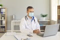 Doctor in face mask having video call with patient sitting at desk with laptop in hospital office Royalty Free Stock Photo