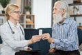 Doctor and geriatric patient at home. Pleasant friendly blond woman doctor giving pills to senior bearded man patient Royalty Free Stock Photo