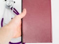 Doctor holding in hand red medical journal, work notebook, purple stethoscope on white back. Close-up Royalty Free Stock Photo