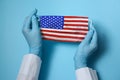 Doctor holding medical mask with USA flag pattern on blue background, closeup. Dangerous virus