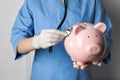 Doctor holding pale pink ceramic piggy bank with stethoscope against white wall, closeup. Medical insurance Royalty Free Stock Photo