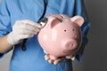 Doctor holding pale pink ceramic piggy bank with stethoscope against white wall, closeup. Medical insurance Royalty Free Stock Photo