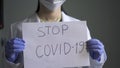 Doctor holds paper sheet with inscription stop covid-19. Close up Shot of female hands wearing rubber gloves on grey