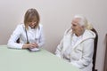 Doctor in a white coat and a stethoscope around her neck, sitting at the table and writing out documents. A very old gray-haired w Royalty Free Stock Photo