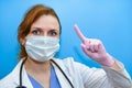 The doctor makes a careful finger - up gesture, close-up. A woman in medical protective gloves and a mask on a blue background,