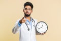 Doctor man showing time on wall office clock, ok, thumb up, approve, pointing finger at camera