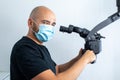 Doctor in medical mask holding handles of black dental microscope and looking aside while working in odontic hospital.