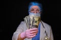 Doctor in medical mask holding money, black background. Nurse with dollars in hands and protective medical gloves, close-up