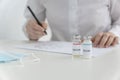 Doctor`s hands hold black pen and writes prescriptions for vaccination, two ampoules with vaccine stand near.