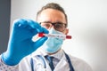 Doctor or scientist with medical mask and glasses holds in hand laboratory test tube with blood, labeled coronavirus, in front of