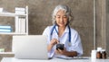 doctor senior grey-haired woman in white medical coat is using a smartphone and smiling while sit use laptop at office Royalty Free Stock Photo