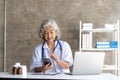 doctor senior grey-haired woman in white medical coat is using a smartphone and smiling while sit use laptop at office Royalty Free Stock Photo