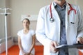 Doctor with stethoscope around neck holding clipboard with documents on background of patient