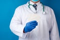 Doctor therapist is dressed in a white robe uniform and blue sterile gloves is standing and holding a stack of empty white paper