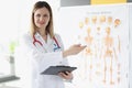 Doctor traumatologist holding medical documents and showing with his hand to poster with human skeleton Royalty Free Stock Photo