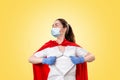 A doctor wearing medical gloves, a surgical mask and a red superhero Cape, tears the medical gown on her chest. Yellow background