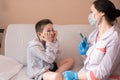 A doctor in a white coat and blue medical gloves holds a syringe with medicine in front of a boy, a child in gray